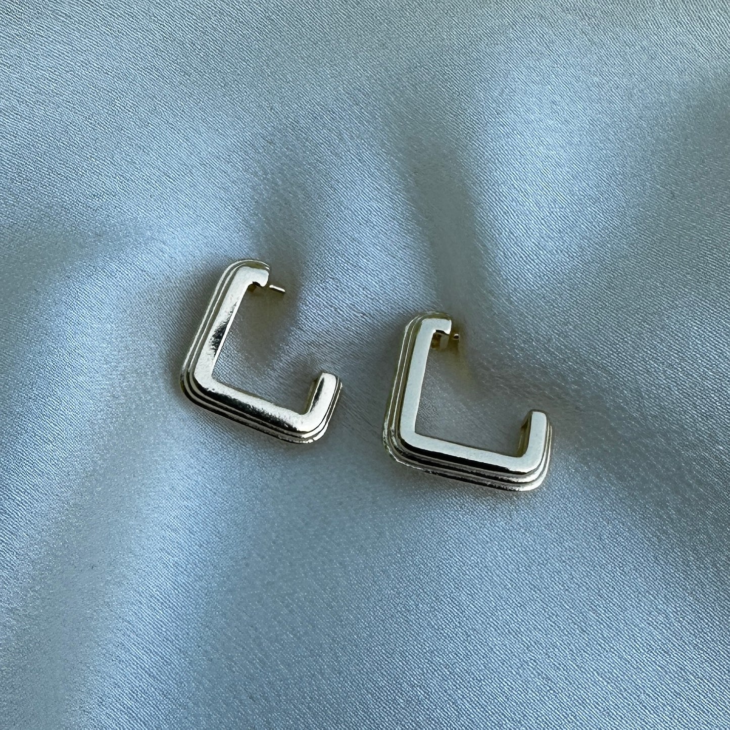 A close up product image of the 18K Gold Filled Grooved C-Hoop Earrings from above angle displayed on white satin fabric