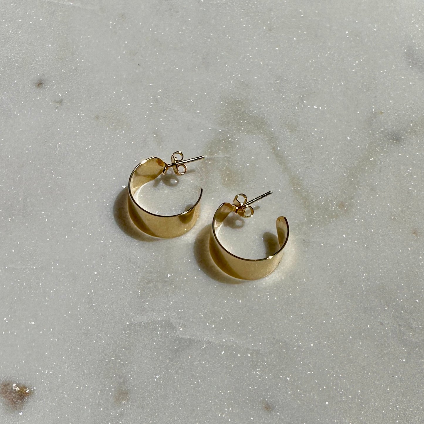 A pair of the 18K Gold Filled Thin Wide Polished Semi Hoop Earrings displayed from above angle on a sparkly surface