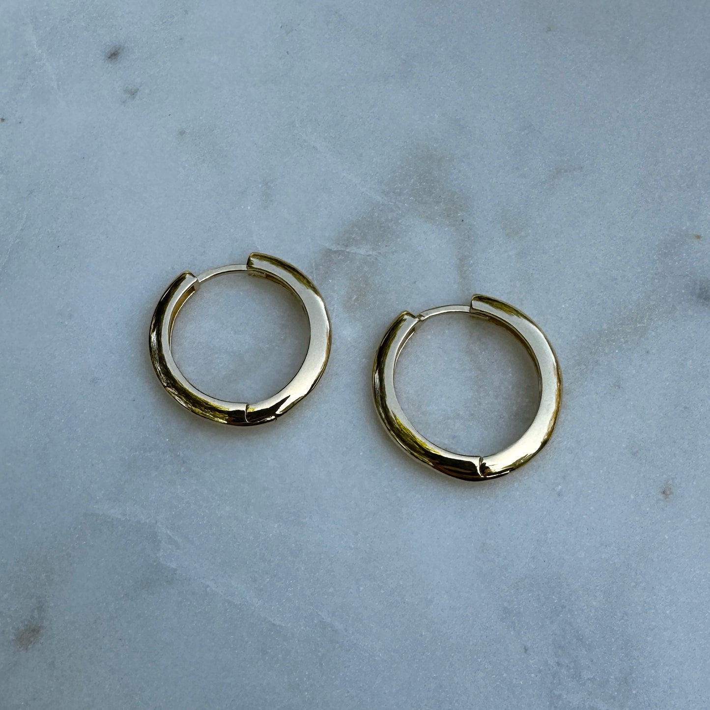 A close up image of a pair of the 18K Gold Filled Lustrous Clicker Hoop Earrings on a marble surface from an above angle