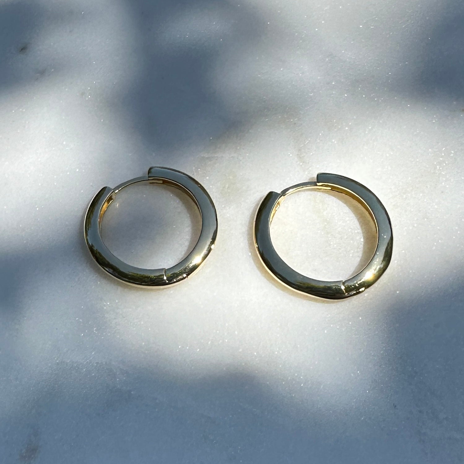 A close up image of a pair of the 18K Gold Filled Lustrous Clicker Hoop Earrings on a marble surface from an above angle with natural light